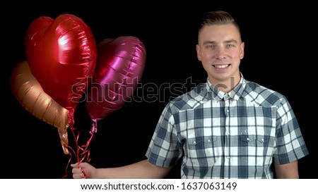 A young man is standing with heart-shaped balloons. Man holds helium balls in his hands on a black background. Valentine's Day is the day of lovers.