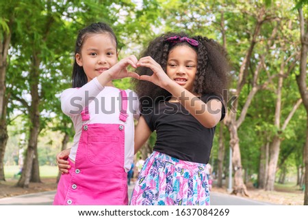 Little cute girl curly hair with her Asian friend making heart shape from hands together while standing in the public park. Cheerful girl embracing her friend in the summer. Friendship concept. Royalty-Free Stock Photo #1637084269