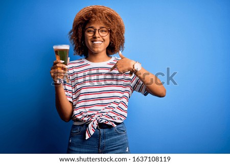 Young beautiful African American afro woman with curly hair on vacation drinking glass of beer with surprise face pointing finger to himself
