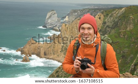 young man in red knitted hat takes picture with modern camera on cliff against azure ocean water and rocks