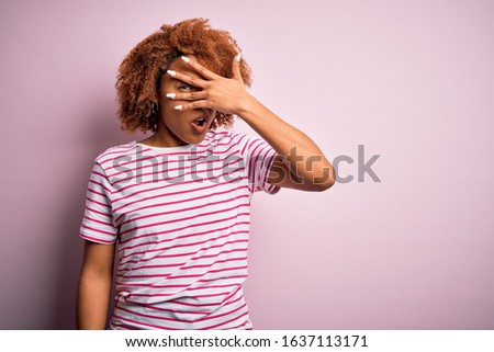 Young beautiful African American afro woman with curly hair wearing casual striped t-shirt peeking in shock covering face and eyes with hand, looking through fingers with embarrassed expression.