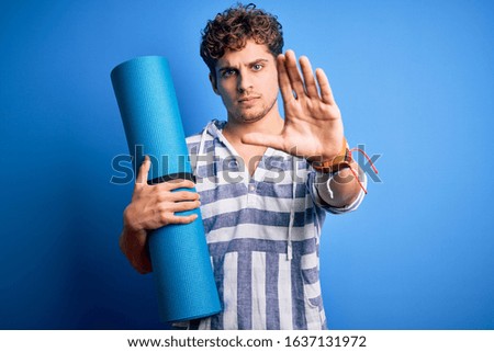 Young blond sporty man with curly hair holding mat to do yoga over isolated blue background with open hand doing stop sign with serious and confident expression, defense gesture