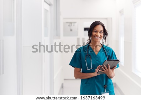 Portrait Of Smiling Female Doctor Wearing Scrubs In Hospital Corridor Holding Digital Tablet Royalty-Free Stock Photo #1637163499