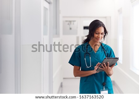 Female Doctor Wearing Scrubs In Hospital Corridor Using Digital Tablet Royalty-Free Stock Photo #1637163520
