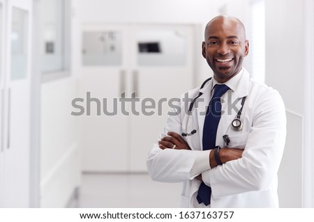 Portrait Of Mature Male Doctor Wearing White Coat Standing In Hospital Corridor Royalty-Free Stock Photo #1637163757