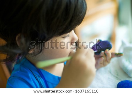 Cute little preschooler painting a rock.Rockpainting activity for homeschooler.Asian boy holding a brush coloring stone.Child with blue shirt with long curly hair.Colorful paint.Fun project for kids.
