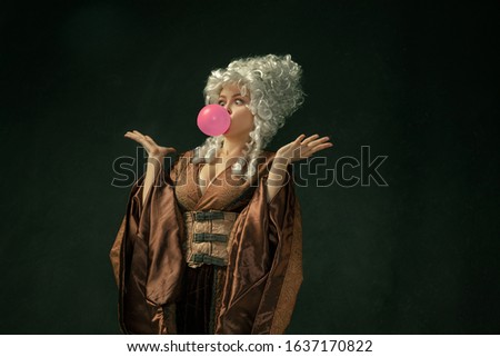 Pink bubble gum. Portrait of medieval young woman in brown vintage clothing on dark background. Female model as a duchess, royal person. Concept of comparison of eras, modern, fashion, beauty.