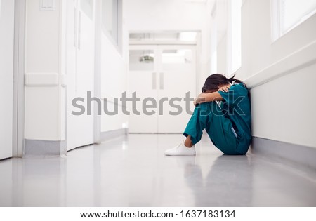 Stressed And Overworked Female Doctor Wearing Scrubs Sitting On Floor In Hospital Corridor Royalty-Free Stock Photo #1637183134