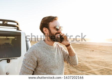 Smiling young man talking on mobile phone while standing at the car at the beach