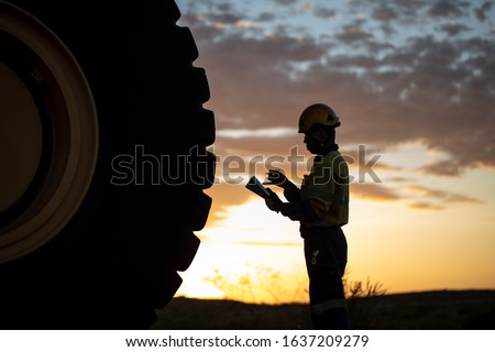 Certified  motor vehicle mechanic mining haul truck driver standing holding inspection pre operation checklist book inspecting taking pictures of defect, damage of wheel tyre sunset background 