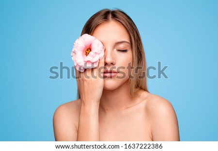 Attractive young woman with clean skin keeping delicate flower near closed eyes while representing spa industry against blue background
