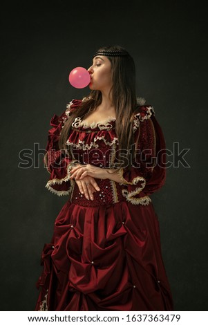 Pink bubble gum. Portrait of medieval young woman in red vintage clothing standing on dark background. Female model as a duchess, royal person. Concept of comparison of eras, modern, fashion, beauty.