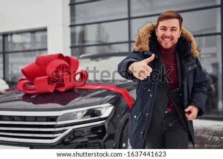 Happy owner of new modern car standing outdoors and smiling. Red gift length on the automobile.