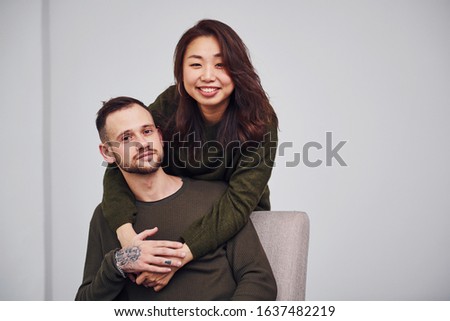 Happy multi ethnic couple in casual clothes embracing each other indoors in the studio. Caucasian guy with asian girlfriend.