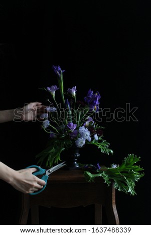 A close up picture of an Asian florist's hands arranging a vase of purple flowers in front of black back ground on a table.