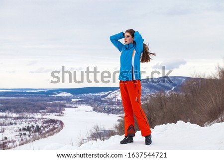 Full-length portrait of a young stylish girl in ski suit posing on a hilltop