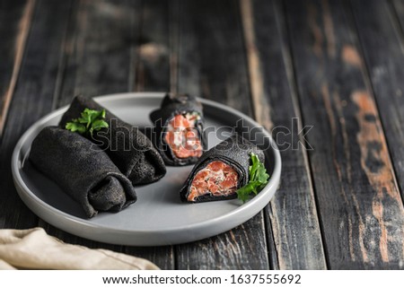 Several black cuttlefish ink pancakes with salmon and cheese filling on a gray plate on a black wooden background. Close up. The Celebration Of Mardi Gras. Horizontal orientation.