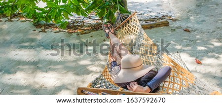 Woman laying on beach swing covering hat on face at beachfront, unrecognized person, sand and tree background in summer holiday, panoramic banner