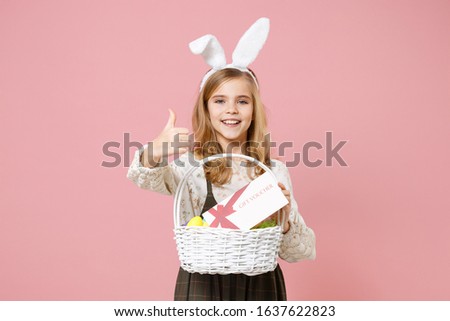 Little pretty blonde kid girl 11-12 years old in spring dress, bunny rabbit ears on head hold in hand wicker basket colorful eggs gift coupon isolated on pastel pink background. Happy Easter concept