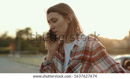 Disappointed young woman talking on the phone standing in the street at road accident. Mixed-up girl in shirt asking for help calling for the police. Car crash.