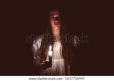 A young scary girl in an old white dress holding a candle in her hand and staring in to the camera. Dark background. Scary horror concept. 