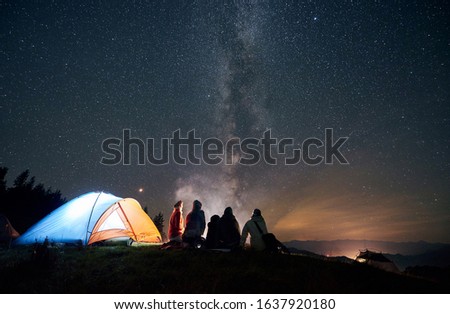 Night summer camping in the mountains. Back view, group of five young friends hikers having a rest together around bonfire beside glowing tourist tent under starry sky full of stars and Milky way.
