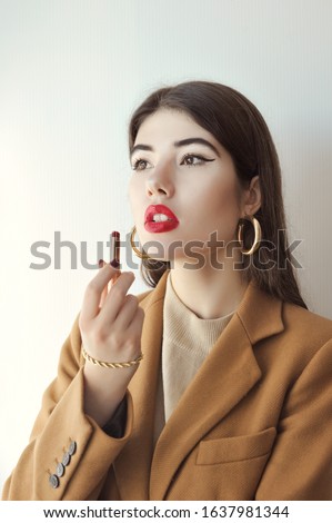 A young brunette girl dressed in a brown jacket with a beige turtleneck and gold fashion jewelry, paints her lips with red lipstick.
