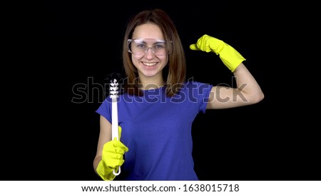A young woman is reaching a toilet with a toilet brush and showing biceps. Woman in safety glasses and gloves with tools for cleaning the toilet. Girl holds a toilet brush. On a black background