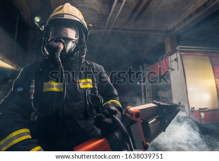 Portrait of a brave firefighter standing confident wearing full protective equipment, turnouts and helmet. Dark background with smoke and blue light.