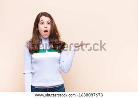 young pretty woman looking surprised and shocked, with jaw dropped holding an object with an open hand on the side against beige background