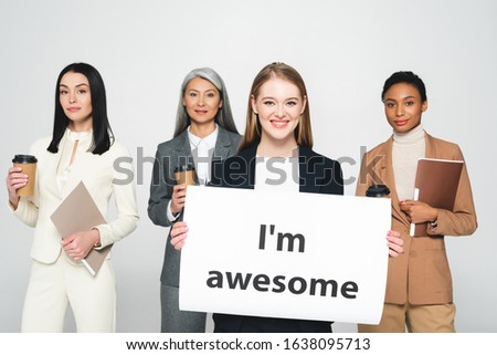 multicultural businesswomen holding folders and disposable cups near placard with i`m awesome lettering isolated on white