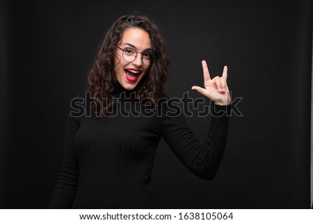 young pretty woman feeling happy, fun, confident, positive and rebellious, making rock or heavy metal sign with hand against black background.