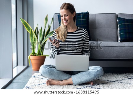Shot of pretty young woman using her mobile phone while working with laptop sitting on the floor at home. Royalty-Free Stock Photo #1638115495