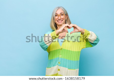 senior or middle age pretty woman smiling and feeling happy, cute, romantic and in love, making heart shape with both hands