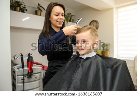 Close up picture of boy/child kid, sitting in the hairdressing salon cutting hair with scissors buy a young female hairdresser.