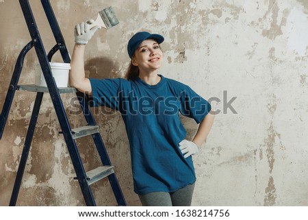 A young woman prepared to do repairs, smiling while standing on the background of a painted wall, in the hands of a brush, next to a ladder.