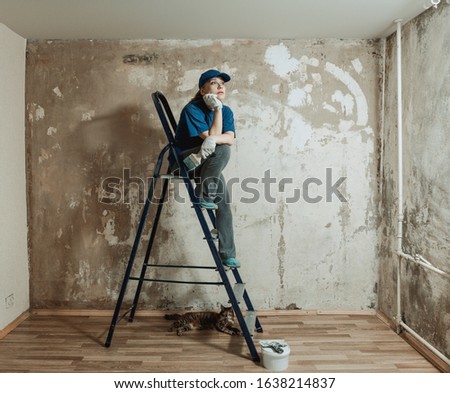 A pensive girl sits on the stairs and looks dreamily in a room with an unpainted wall. DIY repair. Cat under the stairs.