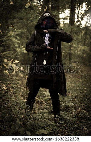 Hooded, face covered wicked sorcerer holding a wolf skull painted in mystic symbols. Mysterious warlock in Twilight of the forest. Halloween witchy atmosphere. Occulture, magic, spiritual, witchcraft.