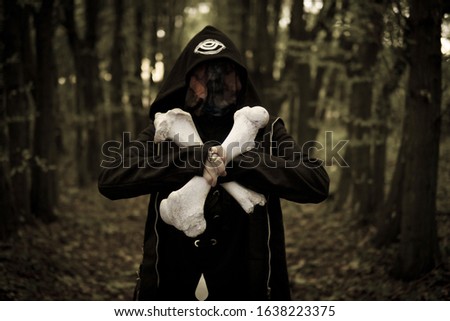 Hooded, face covered, wicked sorcerer holding two big animal bones crossed together. Mysterious warlock in Twilight of the forest. Halloween witchy atmosphere. Occulture, magic, spiritual, witchcraft.