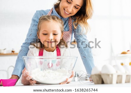 Selective focus of smiling mother and daughter near bowl with flour at kitchen table