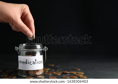 Woman putting coin into glass jar with tag MEDICATION on black table, closeup. Space for text