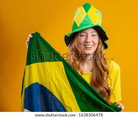 Brazil supporter. Brazilian redhead woman fan celebrating on soccer, football match on yellow background. Brazil colors. Wearing a t-shirt, flag and fan hat.