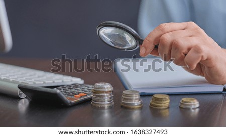 magnifier in hand and stack of coins lies on a dark office desk next to a calculator, pen and keyboard,  concept