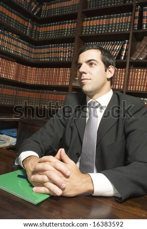 Man in suit sits at desk with hands folded. There are books in the background. Vertically framed photo.