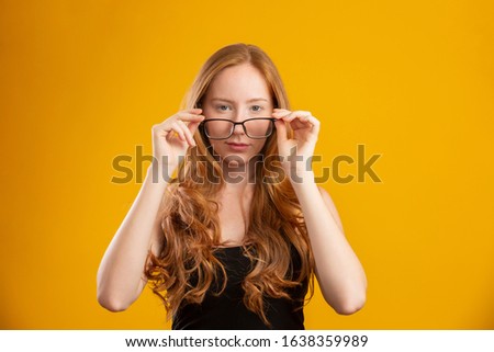Beautiful young redhead woman with curly hair happy with her glasses. Eye care concept. On yellow background.
