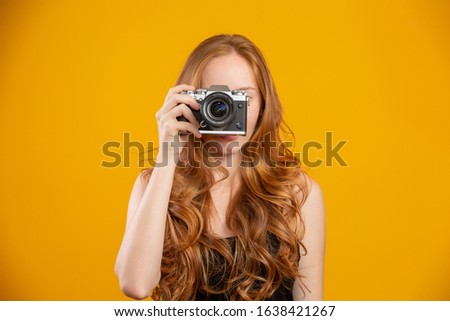 Photo of adorable redhead woman wearing black clothes holding retro vintage camera and taking picture isolated over yellow background. Mock up copy space. Hold retro vintage photo camera.