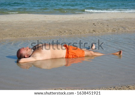 man lying in the puddle. Fat man on the beach. Resort funny pictures. A quiet holiday at the seaside. A man sits in a puddle. Mud bath