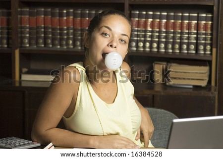 Woman sits at table with lightbulb in her mouth. She is sitting in a library. Horizontally framed photo.