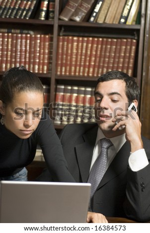 Man in suit talks on cell phone while seated. Woman leans over looking at laptop. Vertically framed photo.