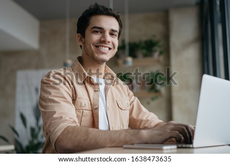 Portrait of happy Indian student studying, learning language. Mixed race business man using laptop computer, typing on keyboard, looking at camera and smiling. Copywriter working from home